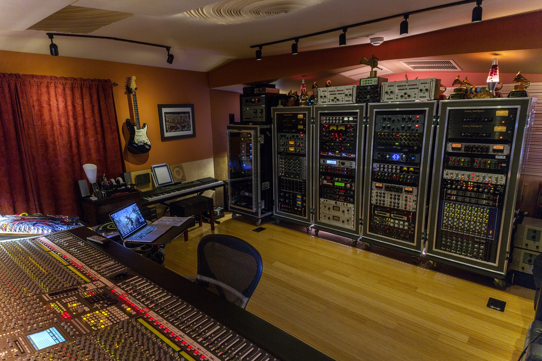 Studio control room with an analog mixing console, a laptop and, on the right, tall racks of outboard gear topped with awards